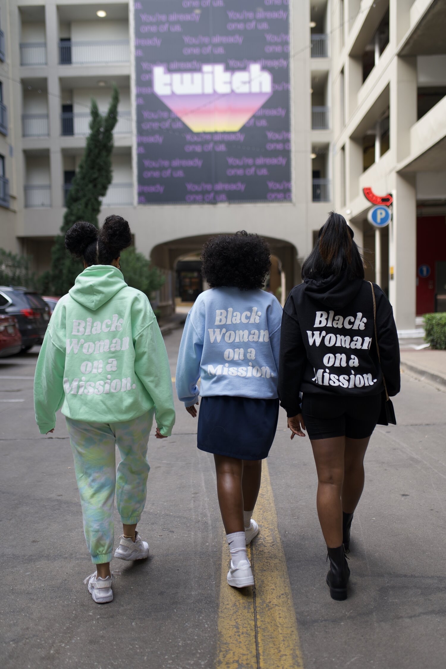 Three women walking with their backs toward the photographer outside with Black Woman On A Mission hoodies and crew necks in various colors,
