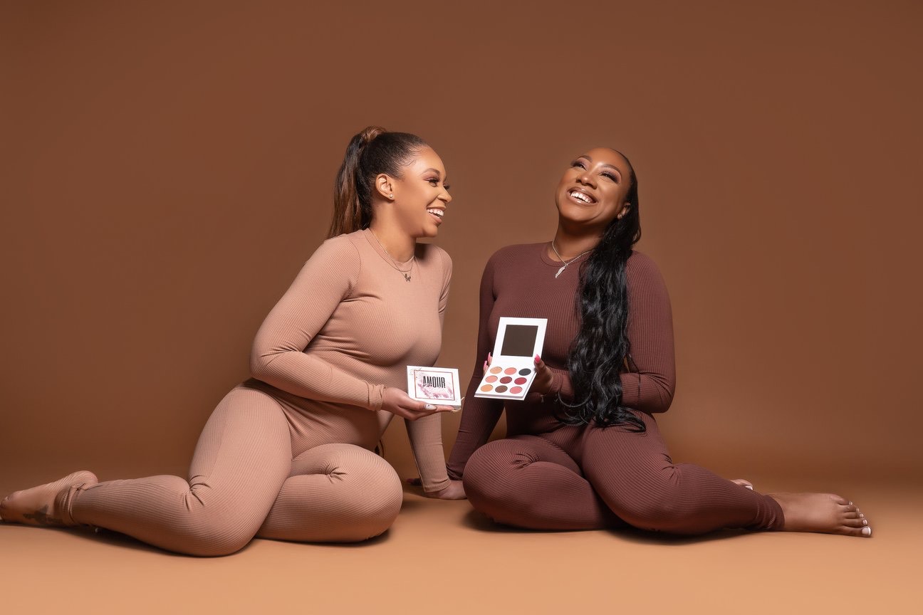 Owners of Beat Cosmetics, Two women sitting on the floor with neutral brown background, holding Beat Cosmetics product.